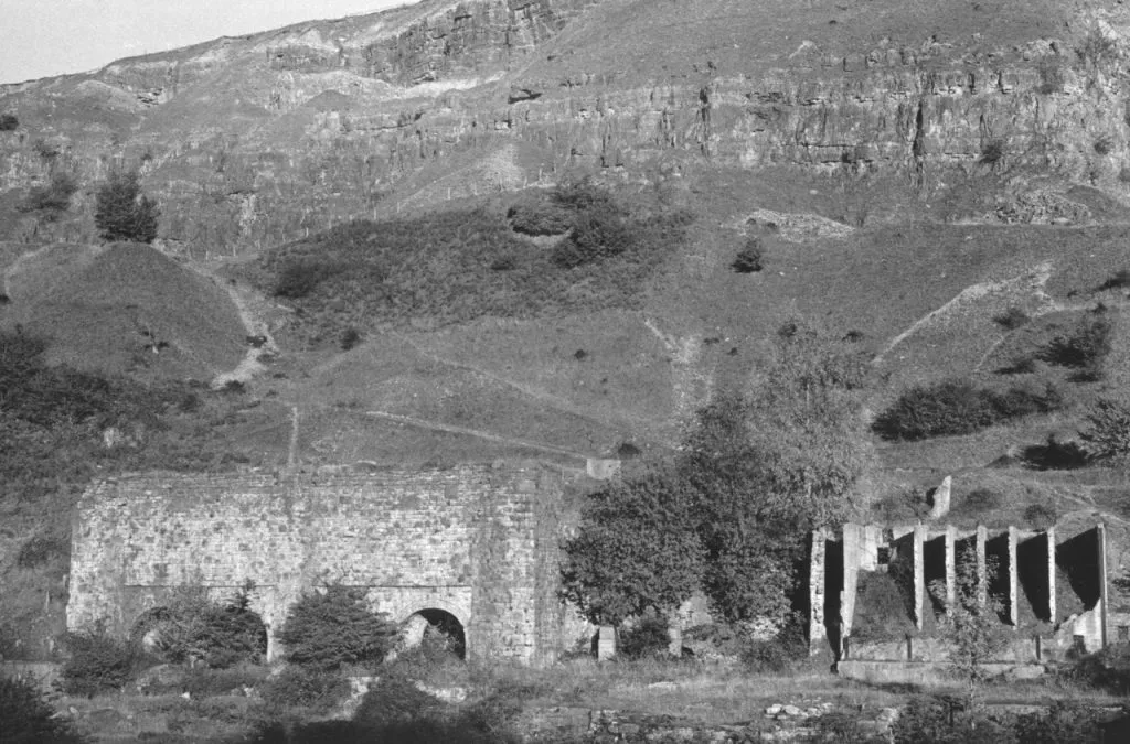 A black and white view of old stone ruins, possibly lime kilns, in the foreground against a steep, rocky, layered hillside or quarry face