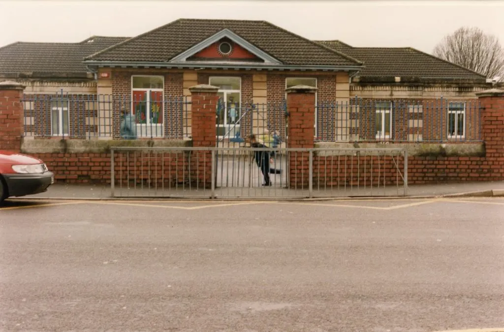 The exterior of a brick school building with a tiled roof, featuring arched windows and a brick and iron fence with an open gate, where a child is running out. A red car is partially visible on the left