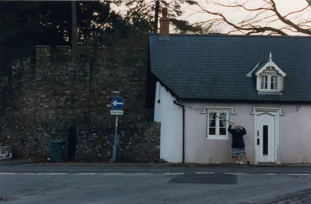 A small white cottage with a dark roof and dormer window next to a tall, ancient stone wall, with a "One Way" sign in the street. A person is leaning against the cottage's window