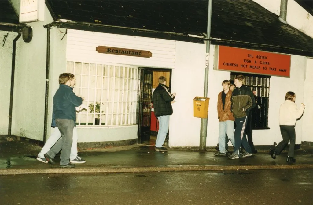 A group of people stand outside a nighttime restaurant called "Restaurant" and "Fish & Chips, Chinese Hot Meals to Take Away", which has white walls and a large, dark red sign