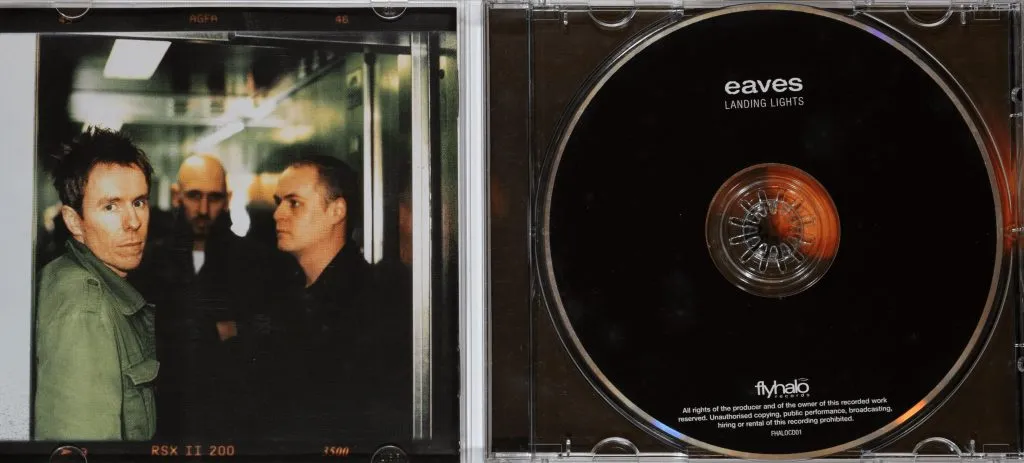 A close-up of the open CD jewel case for the album "Landing Lights" by Eaves, showing a black and white photo insert of three men on the left, and the plain black CD labelled "eaves LANDING LIGHTS" on the right