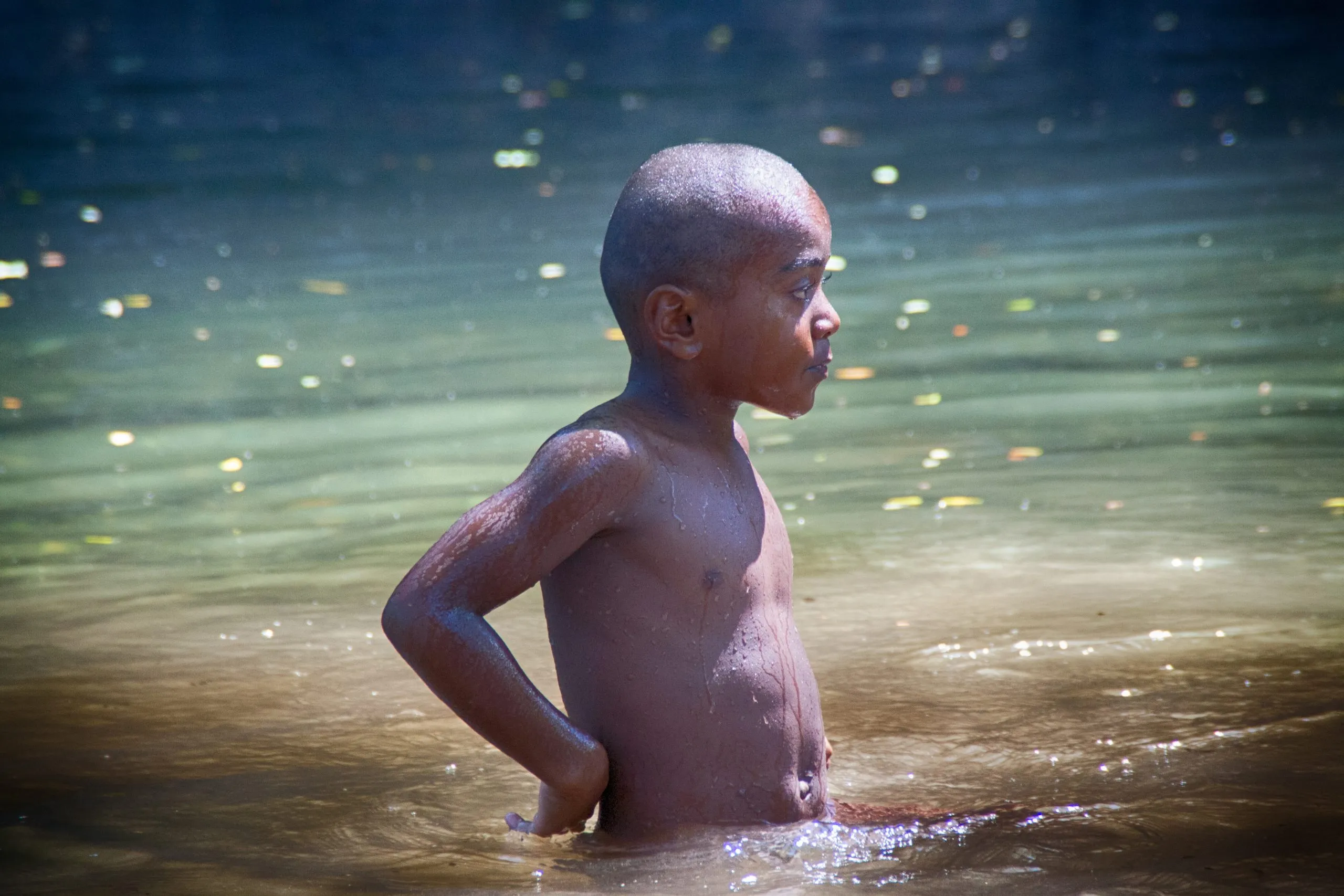 A young, shirtless boy with a shaved head stands waist-deep in murky, sun-dappled water, looking off to the side