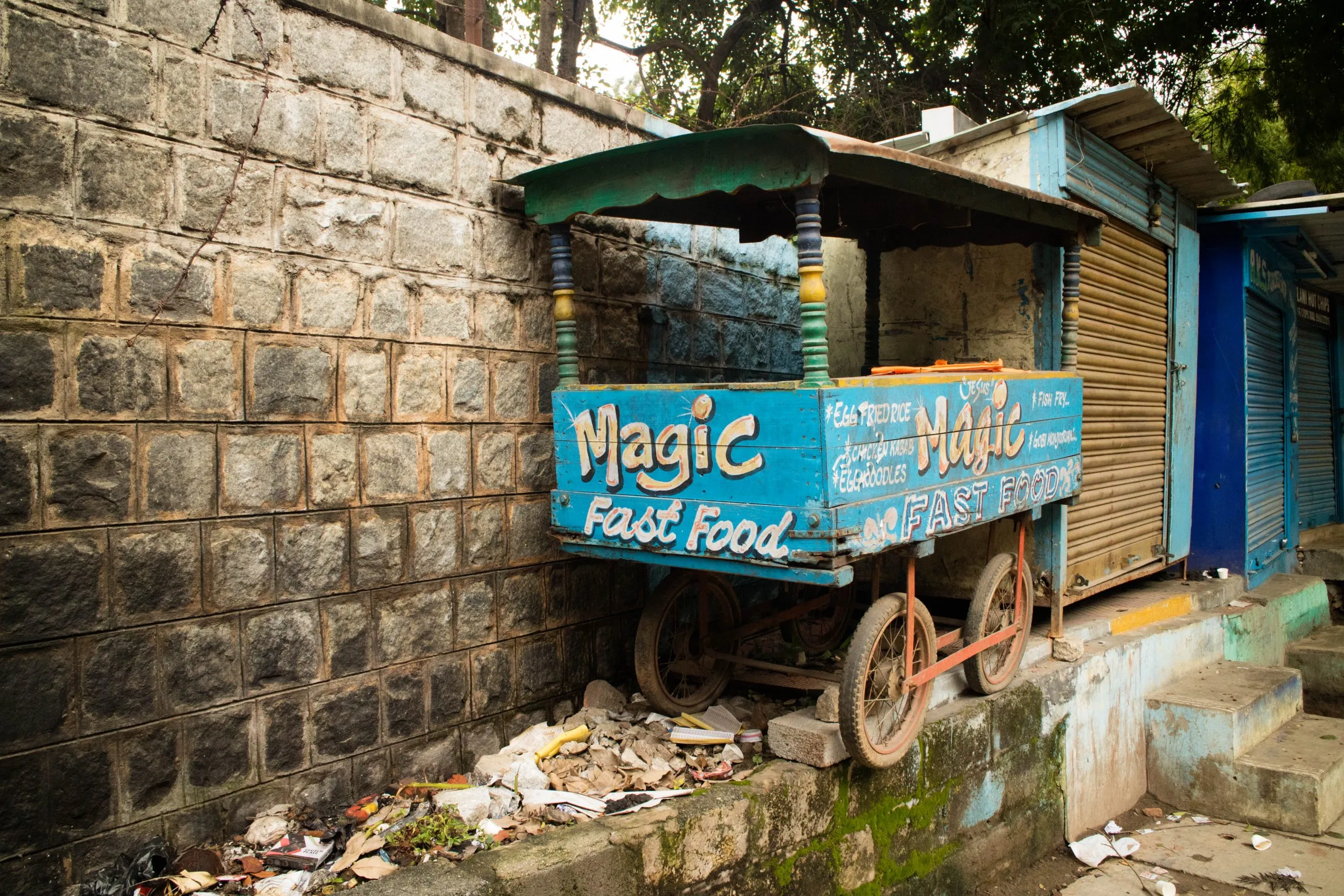 A brightly painted, dilapidated turquoise street cart labelled "Magic Fast Food" rests against a rough stone wall, surrounded by litter and old debris