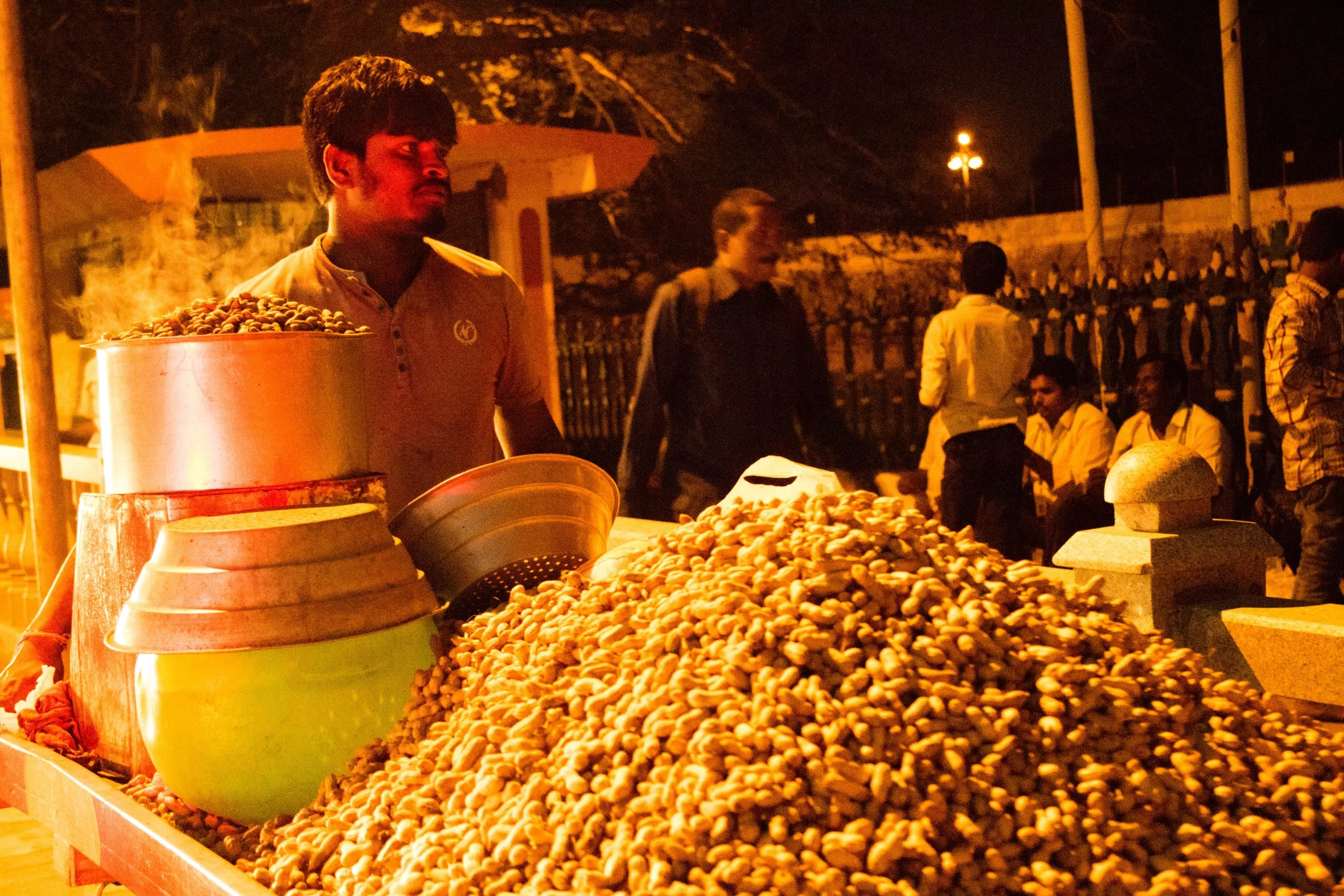 A vendor, illuminated by warm orange light at night, stands behind a large pile of steamed peanuts in their shells next to metal pots, with other people blurred in the background