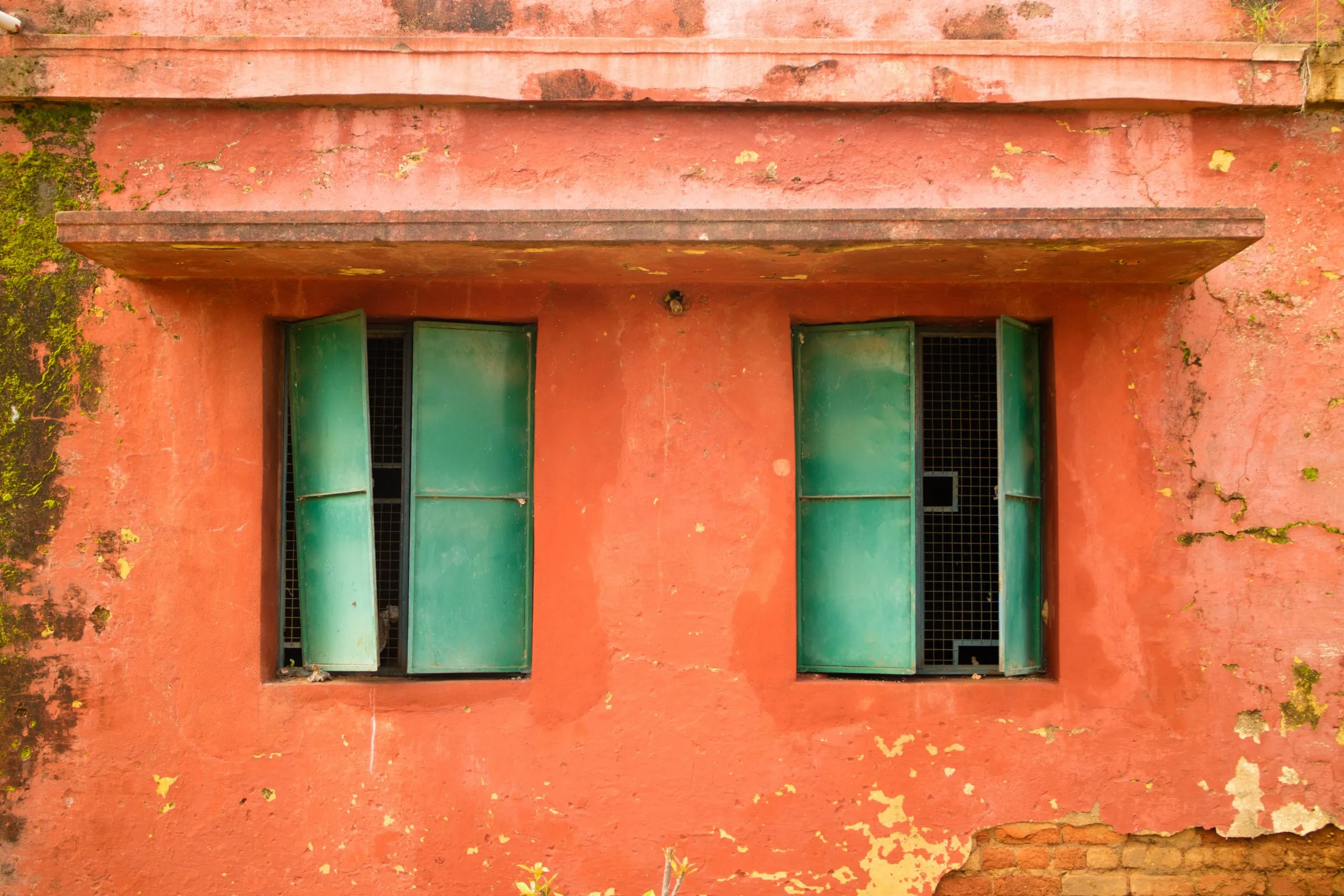 Two bright green, slightly ajar window shutters contrast sharply with a weathered, textured reddish-orange wall with peeling paint and moss
