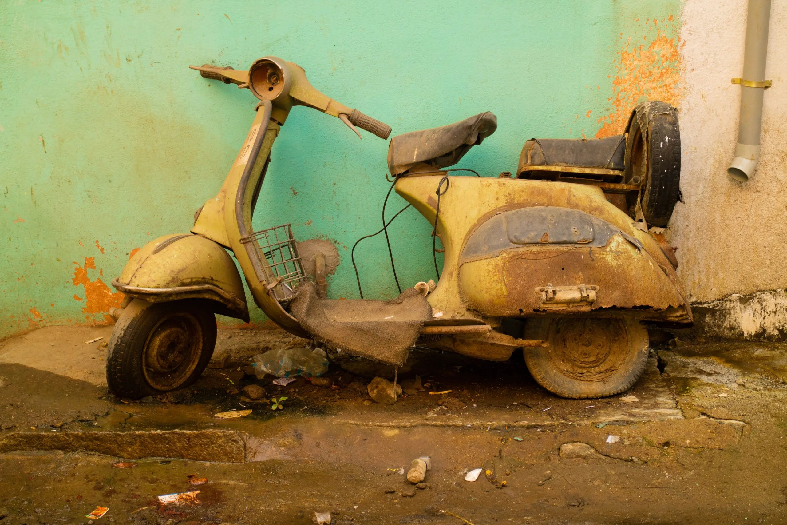 A severely rusted and dirty vintage Vespa scooter is propped up against a peeling light-green wall on a muddy, rubbish-strewn patch of ground