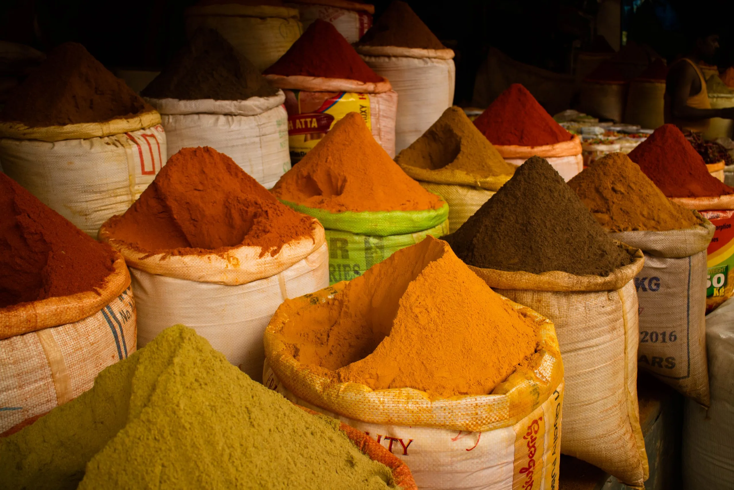 A colourful display of spices in large, open sacks, piled high in cones of bright orange, deep red, yellow, and brown powders at a market stall