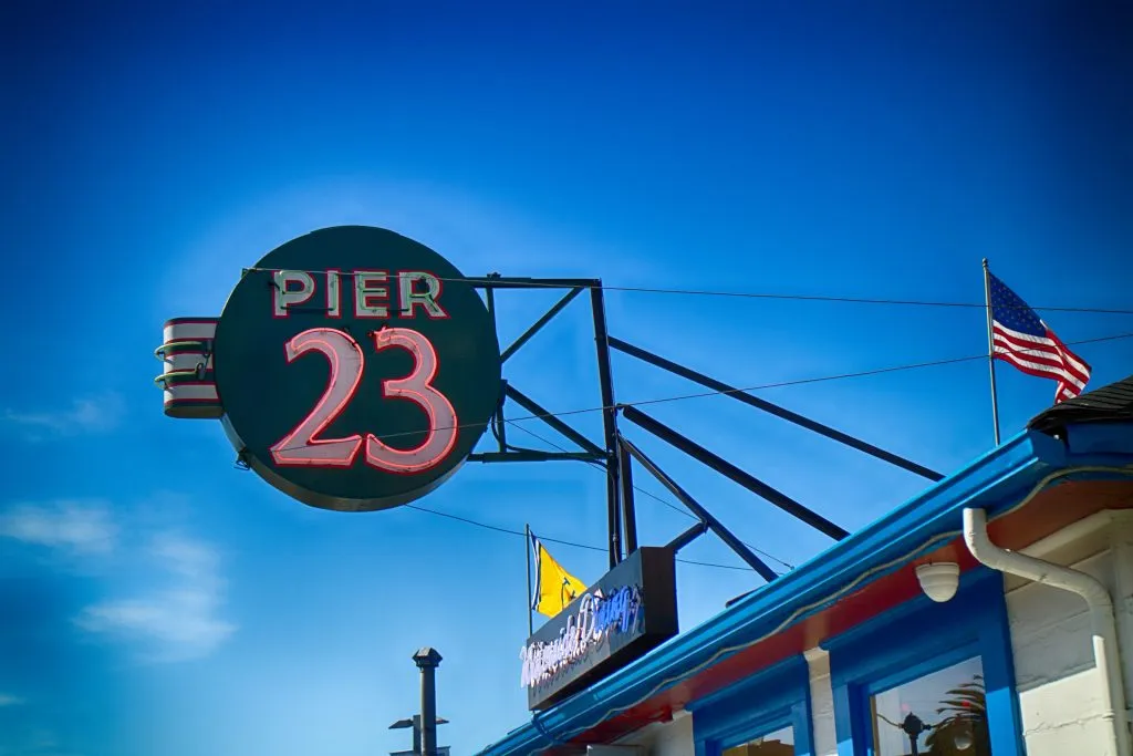 A circular sign for "PIER 23" with bright pink neon lettering is mounted on a steel structure against a clear blue sky, next to a building with an American flag