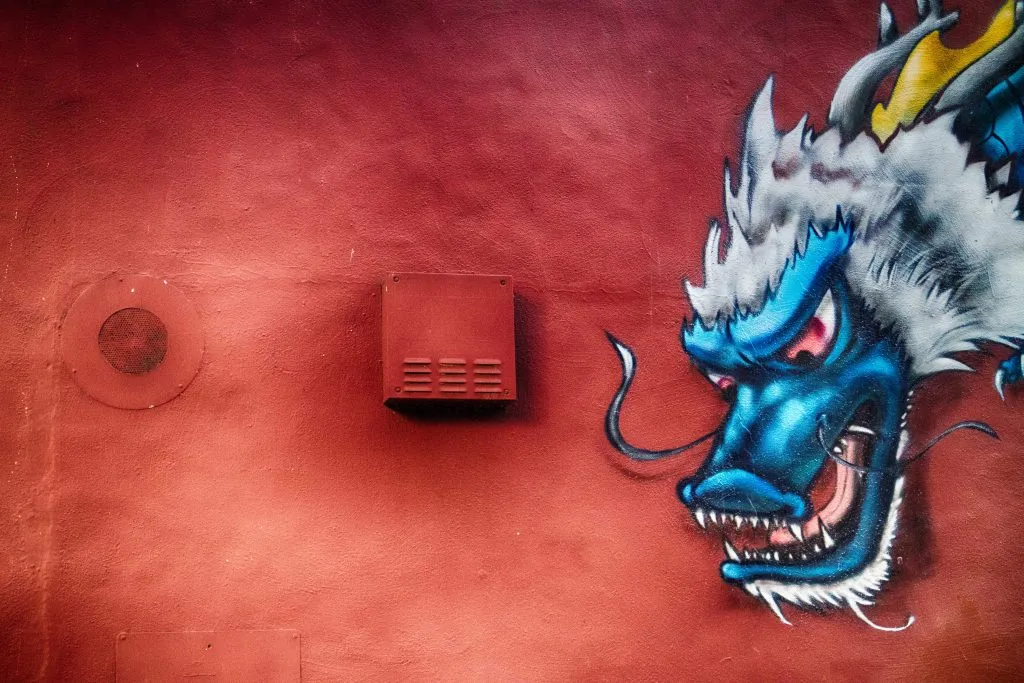A close-up of a vibrant blue and white dragon head graffiti mural on a textured red wall, with a small vent box to the left