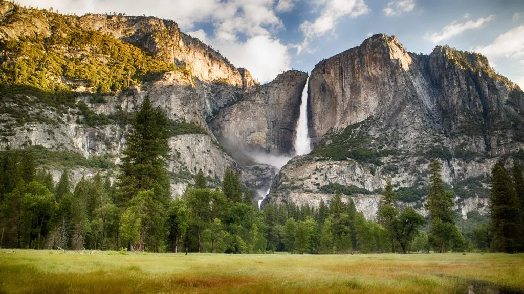 Yosemite Falls cascades down a massive grey cliff face into a lush green meadow and pine forest below, under a bright sun and partly cloudy sky