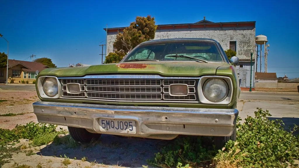 The front view of a weathered, rusted green vintage car (likely a Plymouth Duster) parked on a dirt lot with overgrown weeds in a small town, with a water tower in the distance