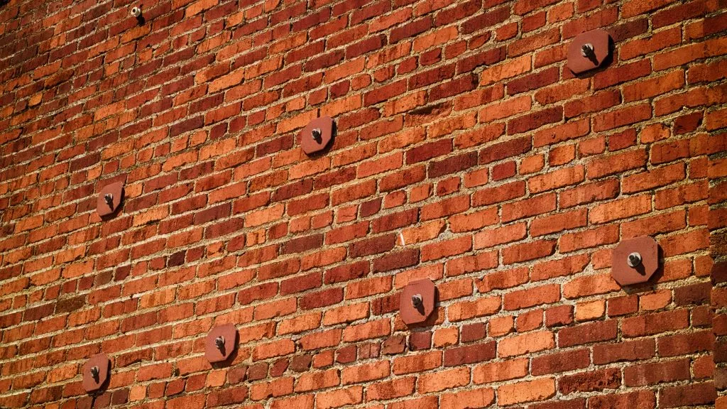 A strong geometric pattern of a red brick wall with several evenly spaced, rusty circular metal plates (earthquake anchors) bolted into it
