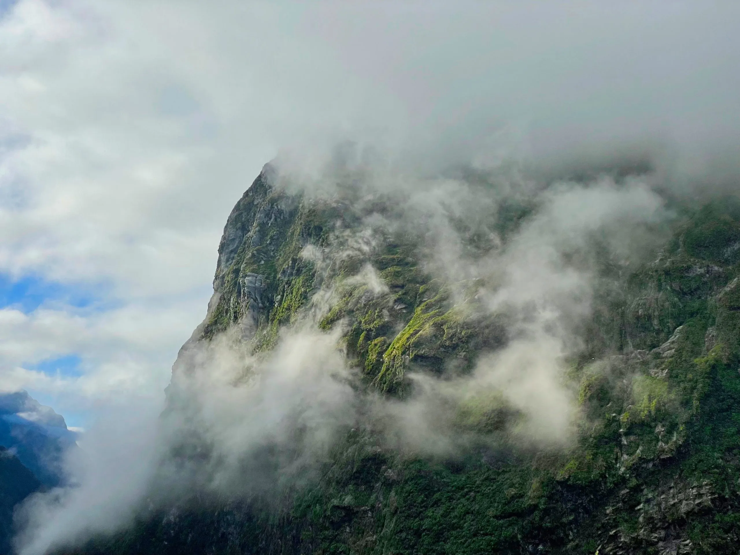 A sheer, mossy green mountain face is partially obscured by thick white mist and low clouds against a partially blue and cloudy sky