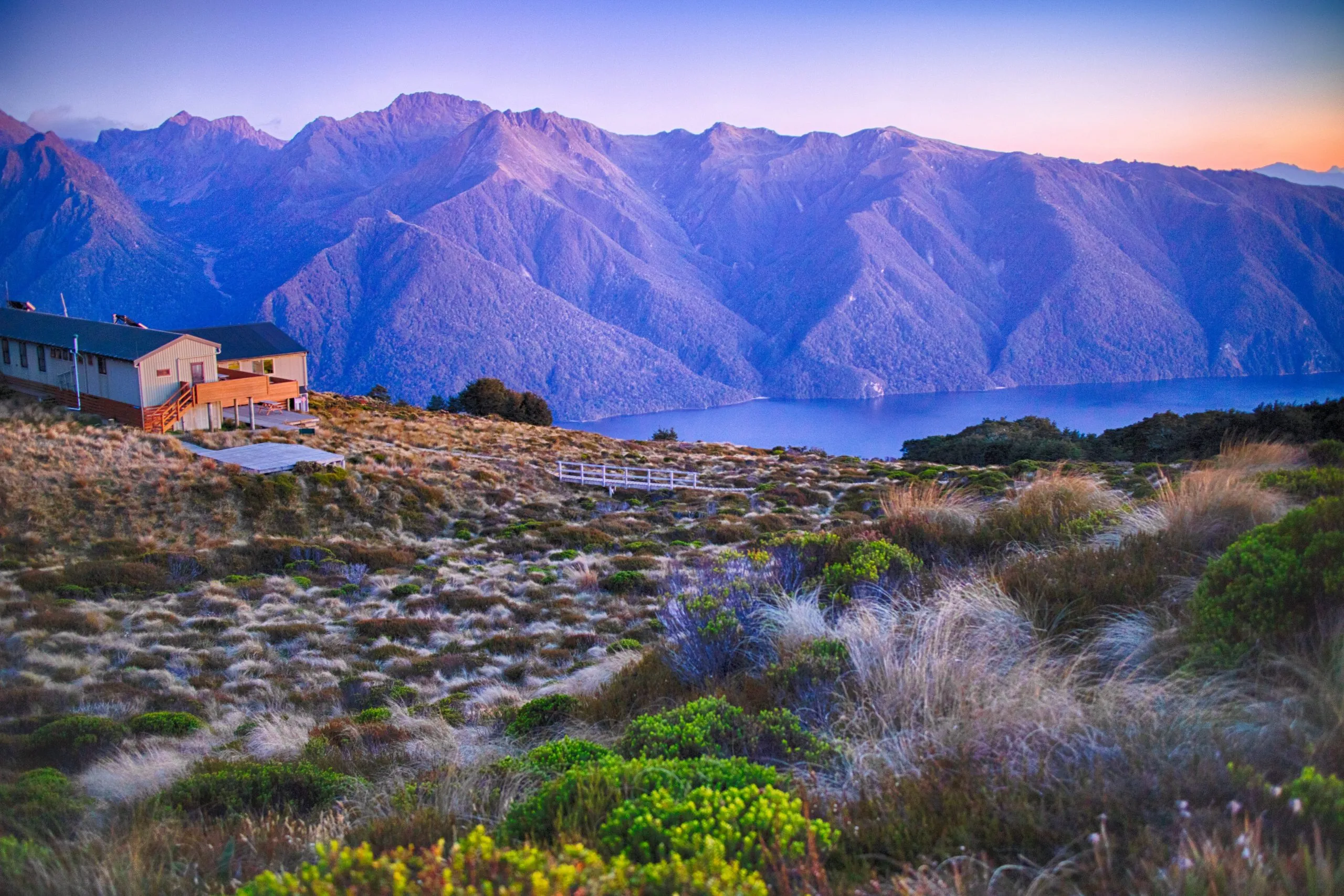A wooden hut or lodge sits on a grassy alpine slope overlooking a deep blue lake with massive, purple-hued mountains rising sharply from the opposite shore at dusk