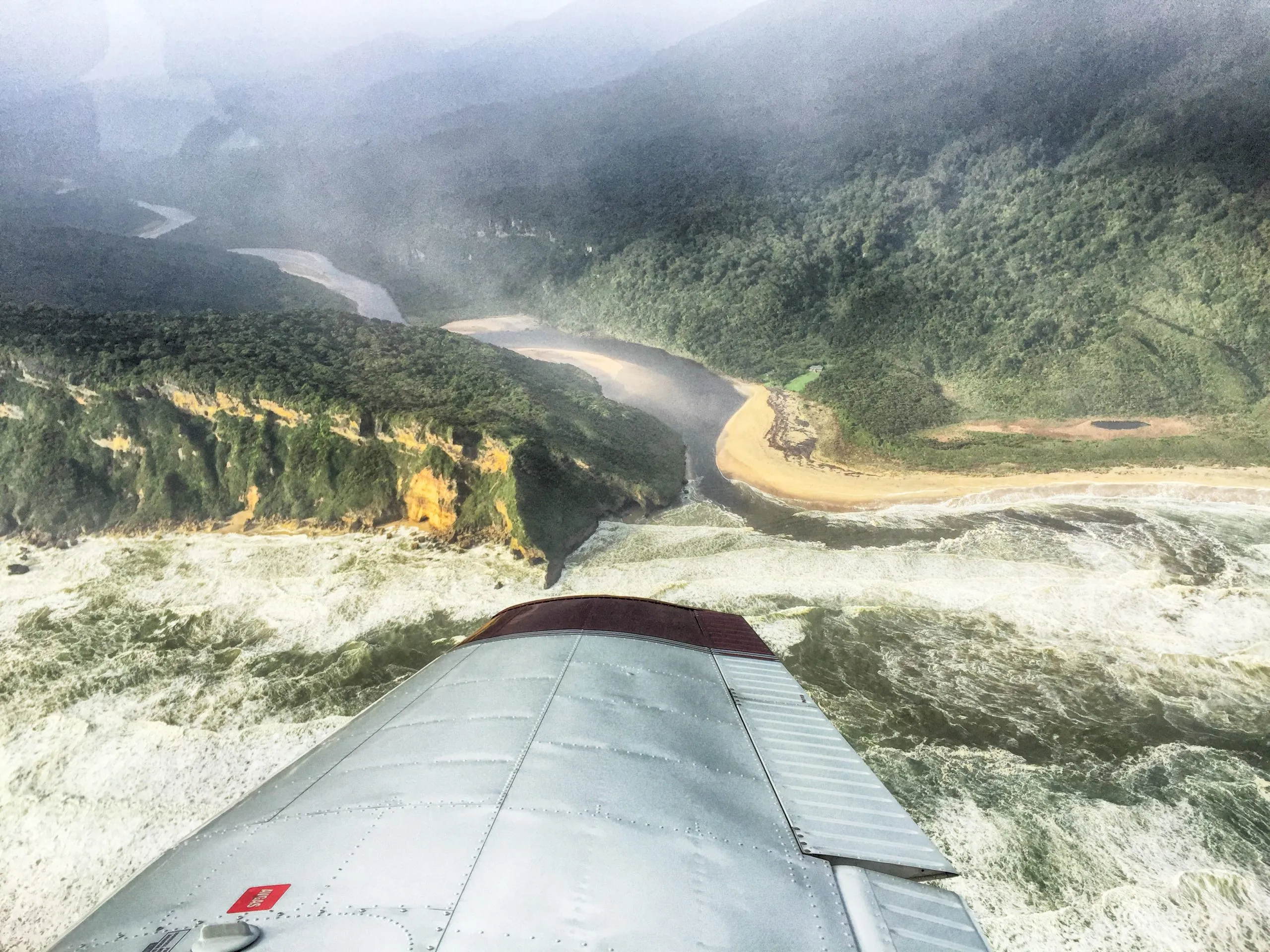 An aerial view from the wing of a plane shows the Heaphy river flowing between high, forested cliffs into the turbulent, white-capped ocean below a misty mountain range