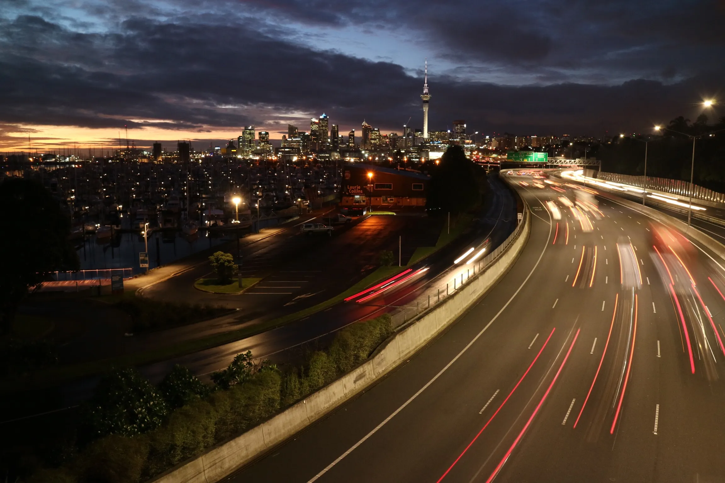 A long exposure photo of the Auckland skyline at night, showing the Sky Tower illuminated, a busy highway with red and white light trails, and a marina full of docked boats
