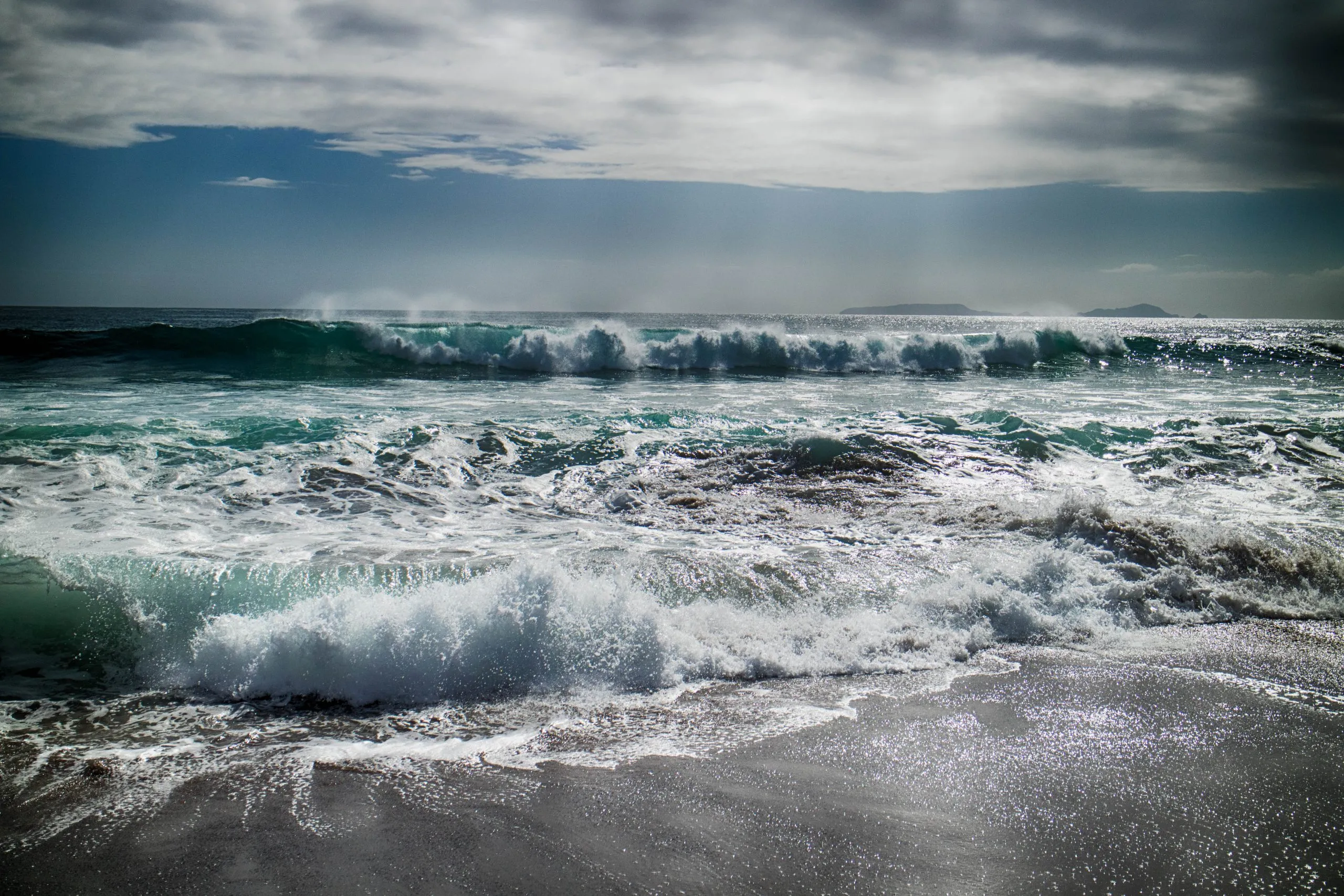 Turquoise ocean waves crash onto a dark, wet sand beach under a dramatic, cloudy sky, with distant waves visible on the horizon