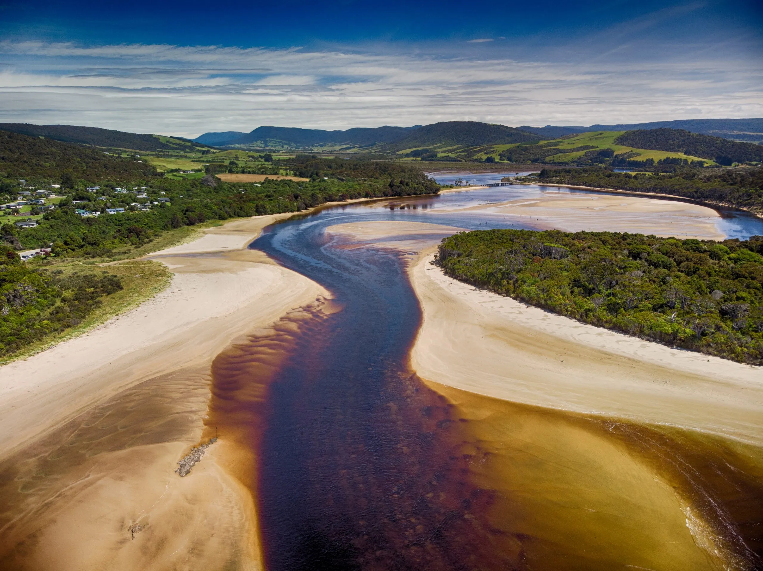 An aerial view shows a dark river winding through wide, pale brown sandbanks and forest, with a small coastal town and green rolling hills in the background under a blue sky