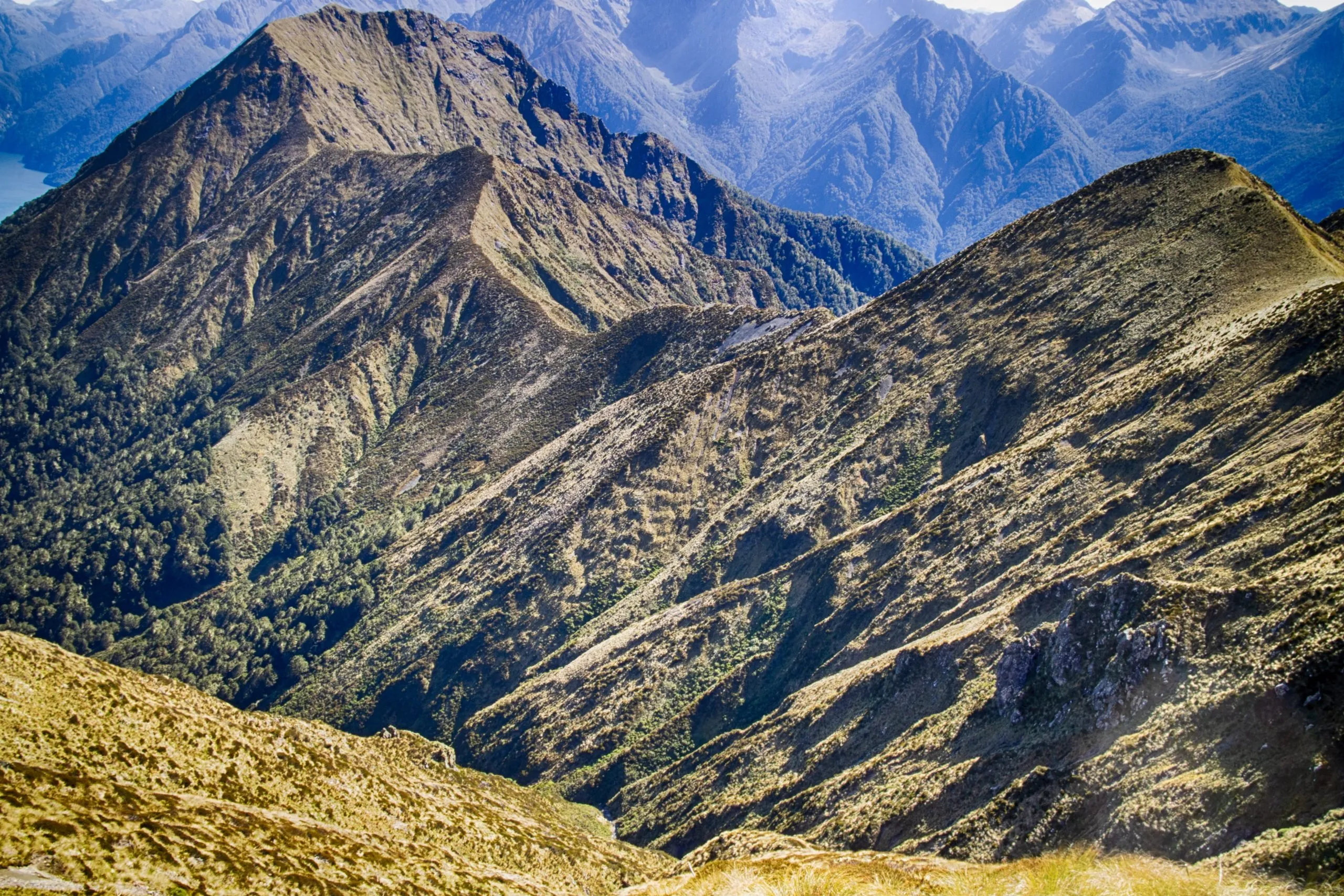 Dramatic, steep alpine ridges covered in low brown and green scrub in the foreground, with layers of blue, hazy mountains receding into the distance