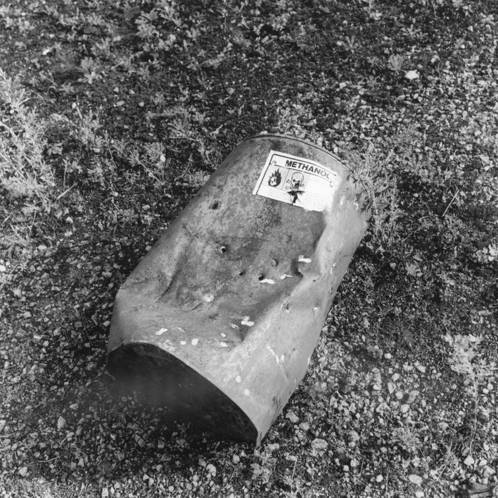A black and white image of a battered, dented metal can with a "METHANOL" warning label, lying on a gravel and dirt ground with small weeds