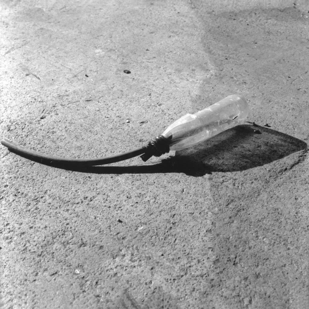A black and white photograph of a clear plastic bottle lying on a rough concrete surface, with a dark rubber tube coming out of the neck, casting a strong shadow